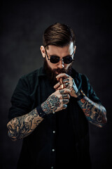 Naklejka premium Portrait of a bearded tattooed guy wearing green shirt and sunglasses lights a cigar