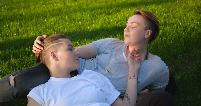 Close up portrait of female same-sex couple lying on the grass. Two pretty stylish girls with short creative hairstyles hug and kiss each other in love.  4k slow motion 50 fps