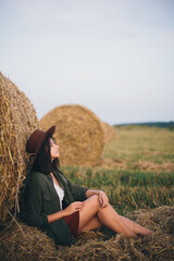Naklejka na ściany i meble Stylish girl relaxing at hay bale in summer field in sunset. Young woman in hat sitting at haystack, atmospheric tranquil moment. Countryside slow life