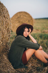 Naklejka na ściany i meble Countryside slow life. Stylish happy girl sitting at hay bale in summer field in sunset. Portrait of young sensual woman in hat smiling at haystack, atmospheric tranquil moment.