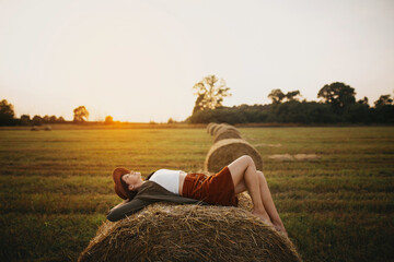 Stylish girl relaxing on hay bale in summer field in sunset. Young woman in hat resting on hay in sunshine, atmospheric tranquil moment. Countryside slow life © sonyachny