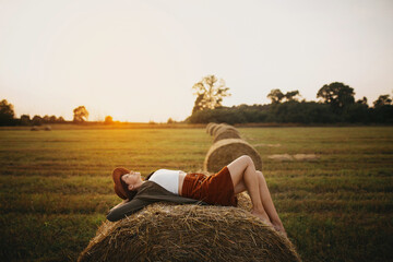 Stylish girl relaxing on hay bale in summer field in sunset. Young woman in hat resting on hay in...