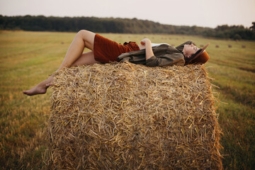 Stylish girl relaxing on hay bale in summer field in sunset. Young woman in hat resting on hay in...