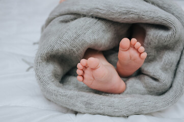 A small child sleeps on a white bed, covered with a gray plaid close-up. The legs of the newborn. Photography, concept.