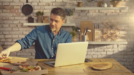 Man working from home on laptop computer, sitting at table in kitchen, eating online ordered pizza. 