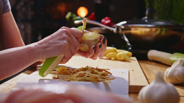 Woman peeling ginger and vegetables for cooking on kitchen table. Closeup hands. Cosy dark room. Real, authentic cooking. 