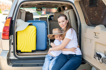 European woman sitting with her son in the trunk of an SUV. Beautiful happy mother loving hugs son. Yellow luggage for summer vacation by car. Holidays with the whole family.