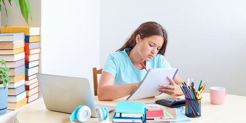 Teenage girl sitting at the table at home with books stationery and laptop and doing assignments in notebook. Studying or learning concept