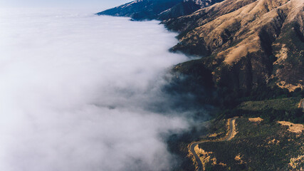 Aerial scenery view of road pass in rocky mountains covered with fog. Bird's eye view through clouds of serpentine asphalt road twists in mountainous terrain on steep slopes