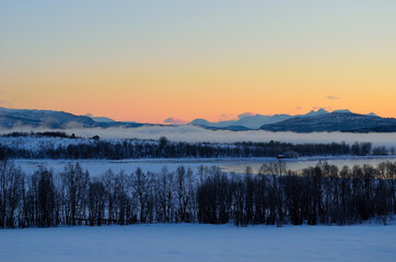 Fototapeta premium ice fog over calm fjord with strong colorful sunset sky and snowy mountain range in winter