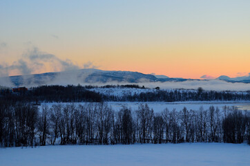 ice fog over calm fjord with strong colorful sunset sky and snowy mountain range in winter