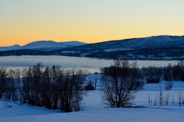 Fototapeta na wymiar ice fog over calm fjord with strong colorful sunset sky and snowy mountain range in winter
