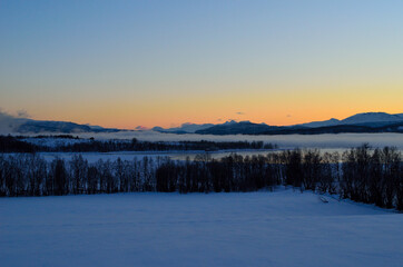 Fototapeta na wymiar ice fog over calm fjord with strong colorful sunset sky and snowy mountain range in winter