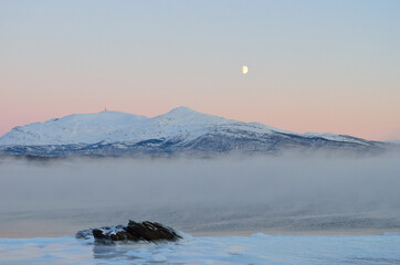 pink sunset sky and full moon over winter fjord with dense ice fog and reflection on water surface