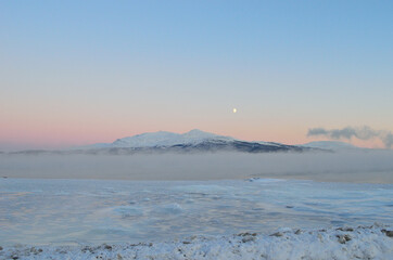 pink sunset sky and full moon over winter fjord with dense ice fog and reflection on water surface