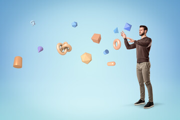 Handsome bearded young man in casual clothes standing and levitating lots of different geometric objects on light blue background.