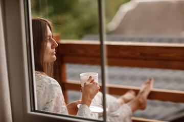 Young female standing after taking a shower in the morning on balcony of the hotel. holding a cup of coffee or tea in her hands. Looking outside nature forest and Mountain