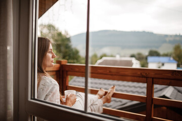 Young female standing after taking a shower in the morning on balcony of the hotel. holding a cup of coffee or tea in her hands. Looking outside nature forest and Mountain