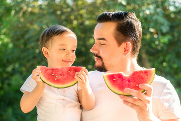 Father and son together eating watermelons at picnic, both holding slices of watermelons, drops of juice on faces and shirts. Dad and kid are looking at each other. Green trees in background