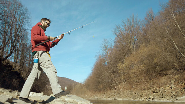 Adult man fishing at mountain stream. Throws a fishing rod into the river at the rocky terrain. Rolling the reel with fishing line.