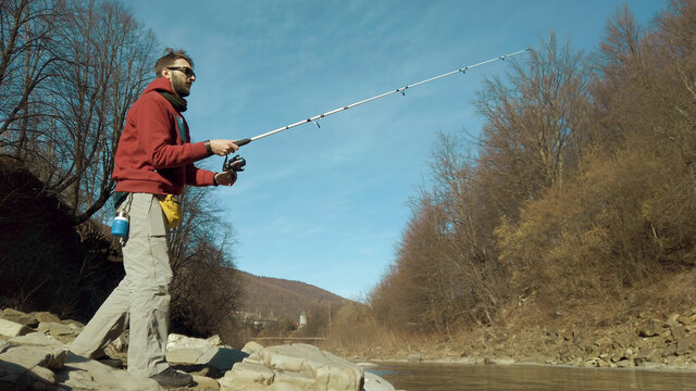 Adult man fishing at mountain stream. Throws a fishing rod into the river at the rocky terrain. Rolling the reel with fishing line.