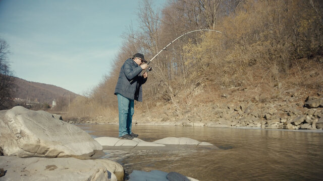 Adult man fishing at mountain stream. Throws a fishing rod into the river at the rocky terrain. Rolling the reel with fishing line.