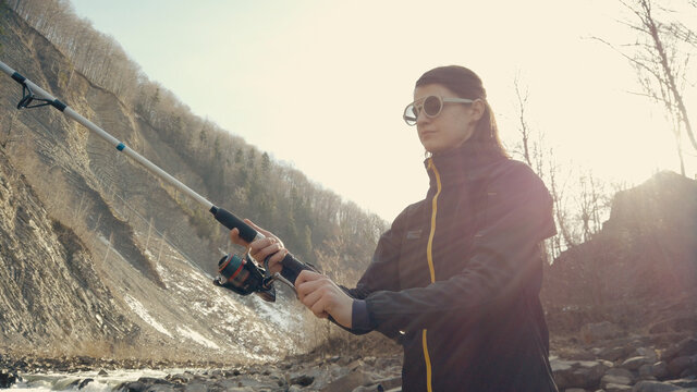 Fisherwoman fishing at mountain stream. Throws a fishing rod into the river at the rocky terrain. Rolling the reel with fishing line. Female fisher. Gender equality