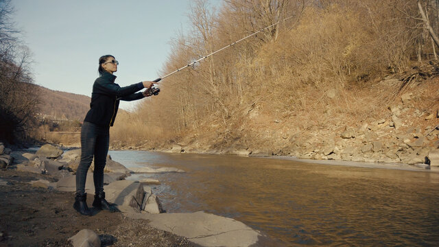 Fisherwoman fishing at mountain stream. Throws a fishing rod into the river at the rocky terrain. Rolling the reel with fishing line. Female fisher. Gender equality