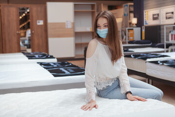Woman in medical mask shopping for new bed during coronavirus quarantine, examining orthopedic...