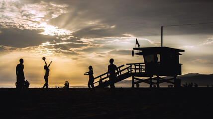 Fototapeta na wymiar Silhouettes of young people playing volleyball by an American beach lifeguard tower by sunset in California