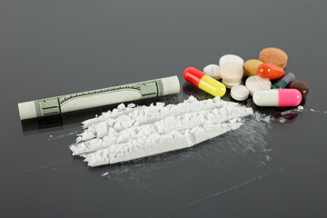 Rolled hundred dollars banknote, cocaine powder and pile of pills on black background.