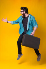 Excited bearded hipster jumping or running with a suitcase over yellow background.