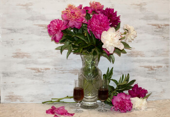 On a table in a crystal vase there is a bouquet of peonies, next to which are two glasses of wine.