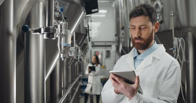 Crop view of man in white lab coat looking at equipment gauge and entering data in tablet. Male brewery worker standing at stainless rows of brewing vats. Female worker at background