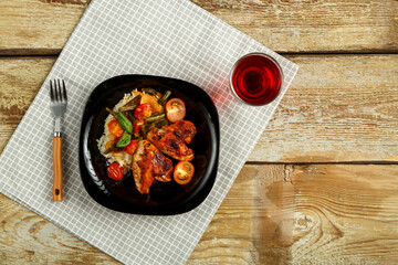Baked chicken wings in tomato sauce with rice and vegetables on a black plate and a glass of red wine on a napkin.