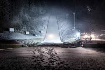 Ski jump at night illuminated by lights covered with snow. Winter sport season. Wide shot, looking up - obrazy, fototapety, plakaty