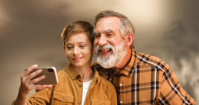 Caucasian teen happy boy taking selfie photo on smartphone camera together with senior grandfather with gray hair and beard. Grandpa and grandson posing to phone and mocking faces. Making funny photos