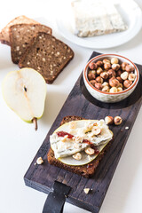 Open sandwich with rye bread, fresh pear , gorgonzola  cheese, hazelnuts and cranberry sauce and bowl with nuts on wooden board and sandwich ingredients  on white background. 