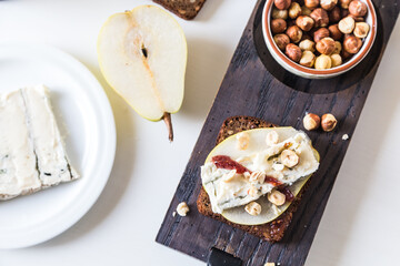 Open sandwich with rye bread, fresh pear , gorgonzola  cheese, hazelnuts and cranberry sauce and bowl with nuts on wooden board and sandwich ingredients  on white background. 