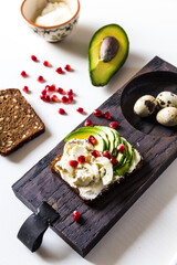 Open sandwich with rye bread, avocado, cream cheese, quail eggs and pomegranate seeds and three quail eggs  on wooden board with sandwich ingredients on white background. 
