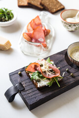 Open sandwich with rye bread, ham, cream cheese, arugula, grated cheese and capers and bowl with mustard sauce on wooden board with sandwich ingredients on white background. 