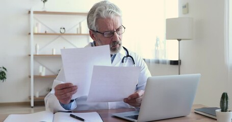 Concentrated elderly senior general practitioner checking paper documents, using computer healthcare software in office. Focused old doctor in white medical coat involved in paperwork in clinic. - Powered by Adobe