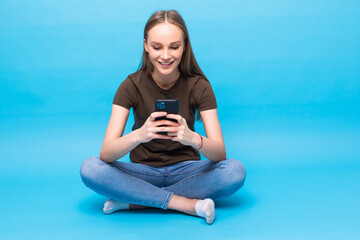 Fototapeta na wymiar Contented smiling woman typing text message or scrolling through social networks using smartphone sitting on the floor isolated over blue background