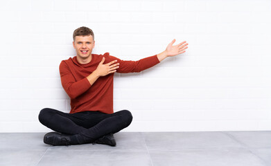 Young handsome man sitting on the floor extending hands to the side for inviting to come