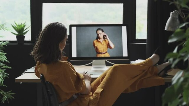 beautiful girl with makeup, dark hair and a yellow linen suit sits in front of a computer putting her feet in shoes on the table and looks at her photos from a photoshoot in the same outfit 