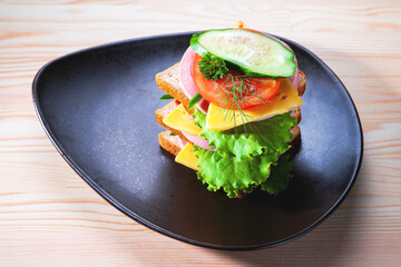 Sandwich with sausage and cheese, herbs, tomato and cucumber on a dark plate of triangular shape. Snack in the afternoon. A sandwich with you. Toasts for breakfast or lunch