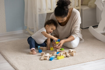 Loving African American young mother and cute toddler girl playing with colorful wooden blocks, sitting on warm floor at home, happy mum and adorable little daughter constructing tower, childcare