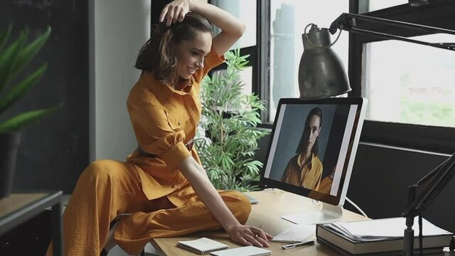 beautiful girl with makeup, dark hair and a yellow linen suit sits on a table in front of a computer in the room and looks at the screen on her photos from a photoshoot in the same outfit