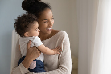 Smiling dreamy African American young mother holding cute little daughter close up, looking to aside at window, dreaming about good future, planning, caring happy mum hugging toddler girl
