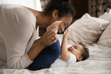 Loving African American mother tickling laughing adorable toddler daughter close up, playing funny game, lying in cozy bed at home, happy young mum and cute little girl having fun together